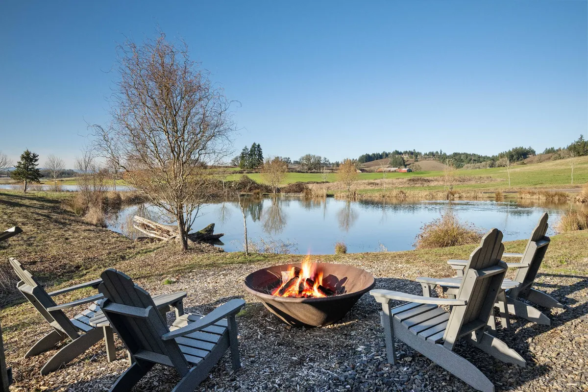 A cozy fire pit surrounded by Adirondack chairs sits by a pond, with expansive views of the surrounding landscape. The peaceful mood invites guests to gather for stories, warmth, and sunset appreciation, reflecting seasonal leisure.