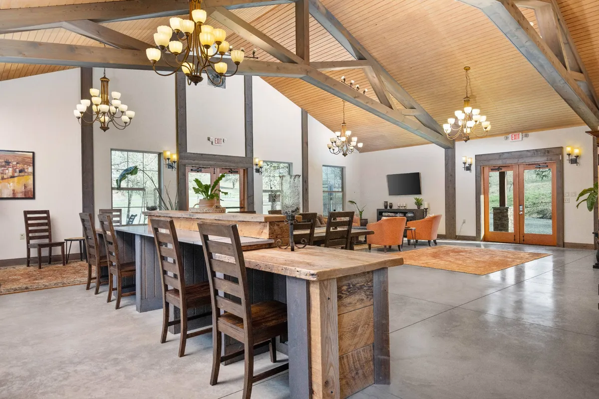 A spacious, open-concept interior featuring a large wooden dining table as the centerpiece, with comfortable seating nearby. Large windows flood the space with natural light and offer glimpses of the scenic outdoors. Elegant chandeliers hang overhead, enhancing the inviting, communal atmosphere.