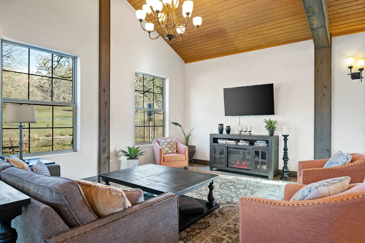 A bright, cozy living room featuring plush sofa seating around a chic coffee table, elegant chandelier overhead, and expansive windows offering serene lake views. The space is perfect for morning coffee, relaxed evenings in, or celebrating special moments with friends or loved ones.
