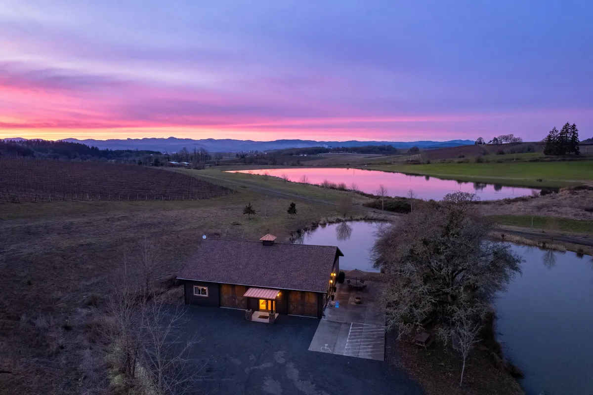 A serene exterior shot highlighting the lakehouse, the sparkling pond, a dramatic sunset, and the vineyard in the background. The scene suggests relaxation after a day of tastings, with opportunities to unwind by the water or explore the vines beyond.