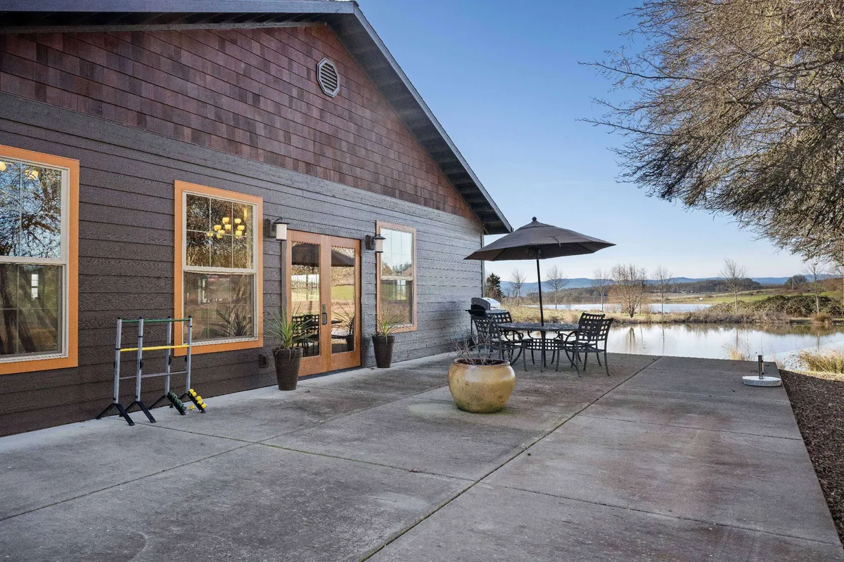 This image features a wooden-sided house with an outdoor dining set and umbrella, surrounded by potted plants, overlooking tranquil water. The scene evokes alfresco dining, relaxation, and intimate gatherings, with a direct connection to the patio experience described in the section.