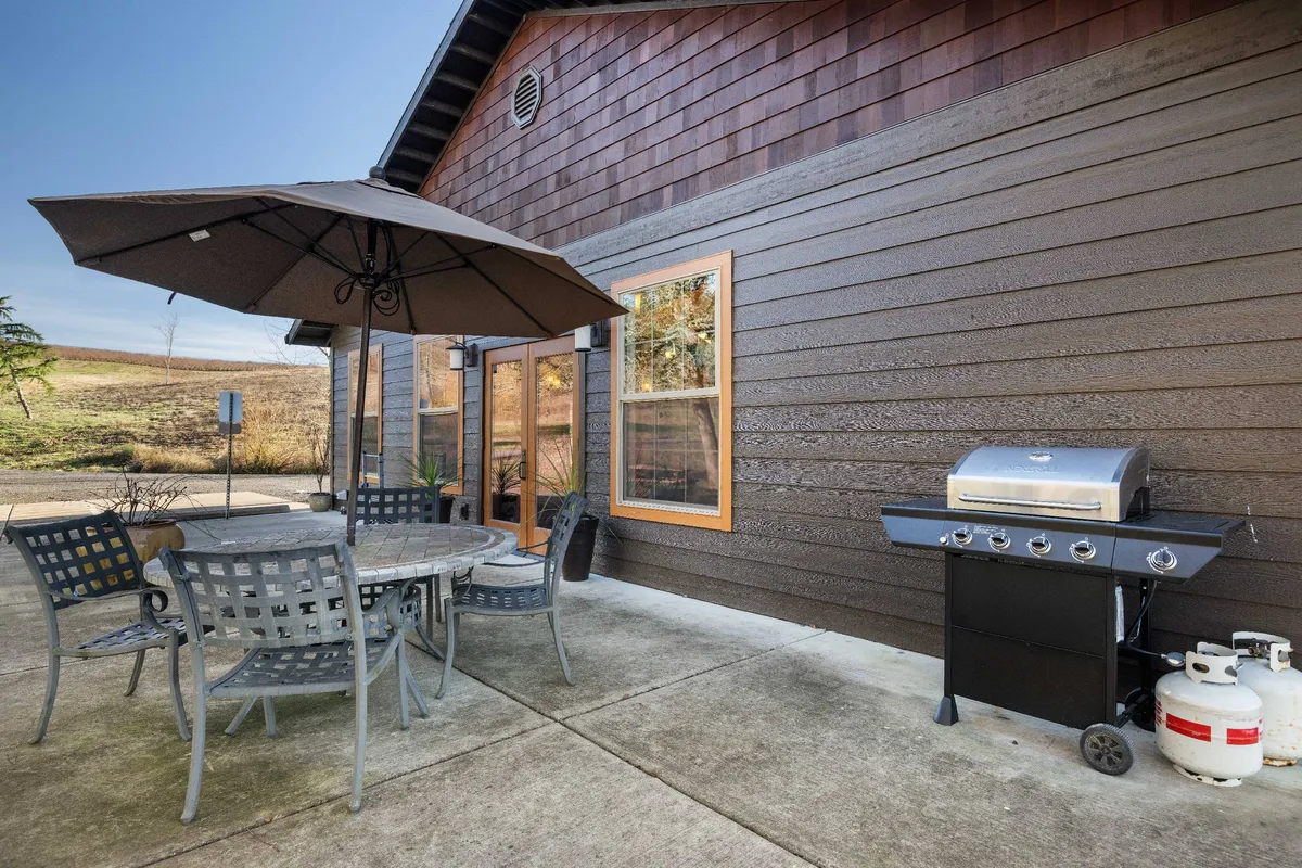 An outdoor dining setup featuring a table shaded by an umbrella, surrounded by chairs and a grill, set against the home’s exterior. The inviting arrangement suggests space for group meals, celebrations, and lively conversation, all with the backdrop of the house and natural landscape.