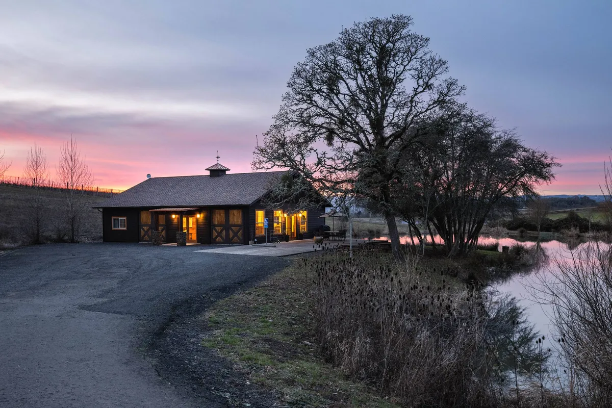 A warm, inviting exterior shot of the Lakehouse just after sunset. The building’s windows glow softly, reflecting the cozy atmosphere inside, while the still water in the foreground and sprawling tree evoke a sense of tranquility. The subtle light in the sky hints at evening activities, making the scene perfect for gatherings by the lake.