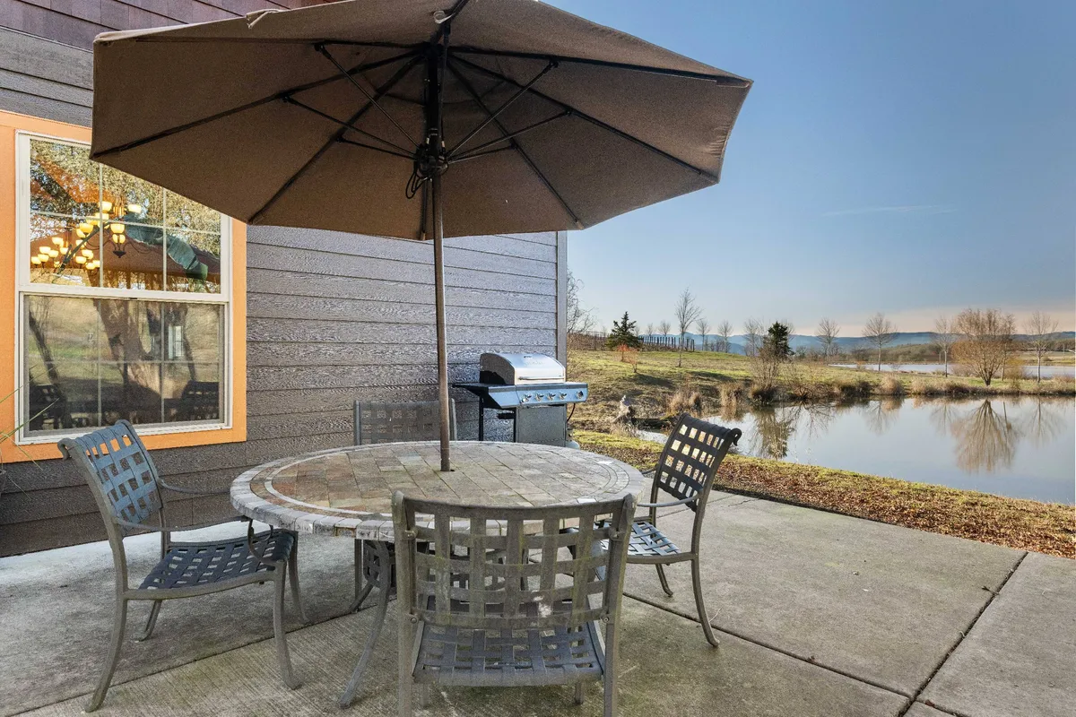 A round outdoor table with an umbrella sits beside a grill, overlooking a peaceful pond. The table is set up for outdoor dining, with the grill indicating barbecue options, and serene water views enhancing the setting's appeal.