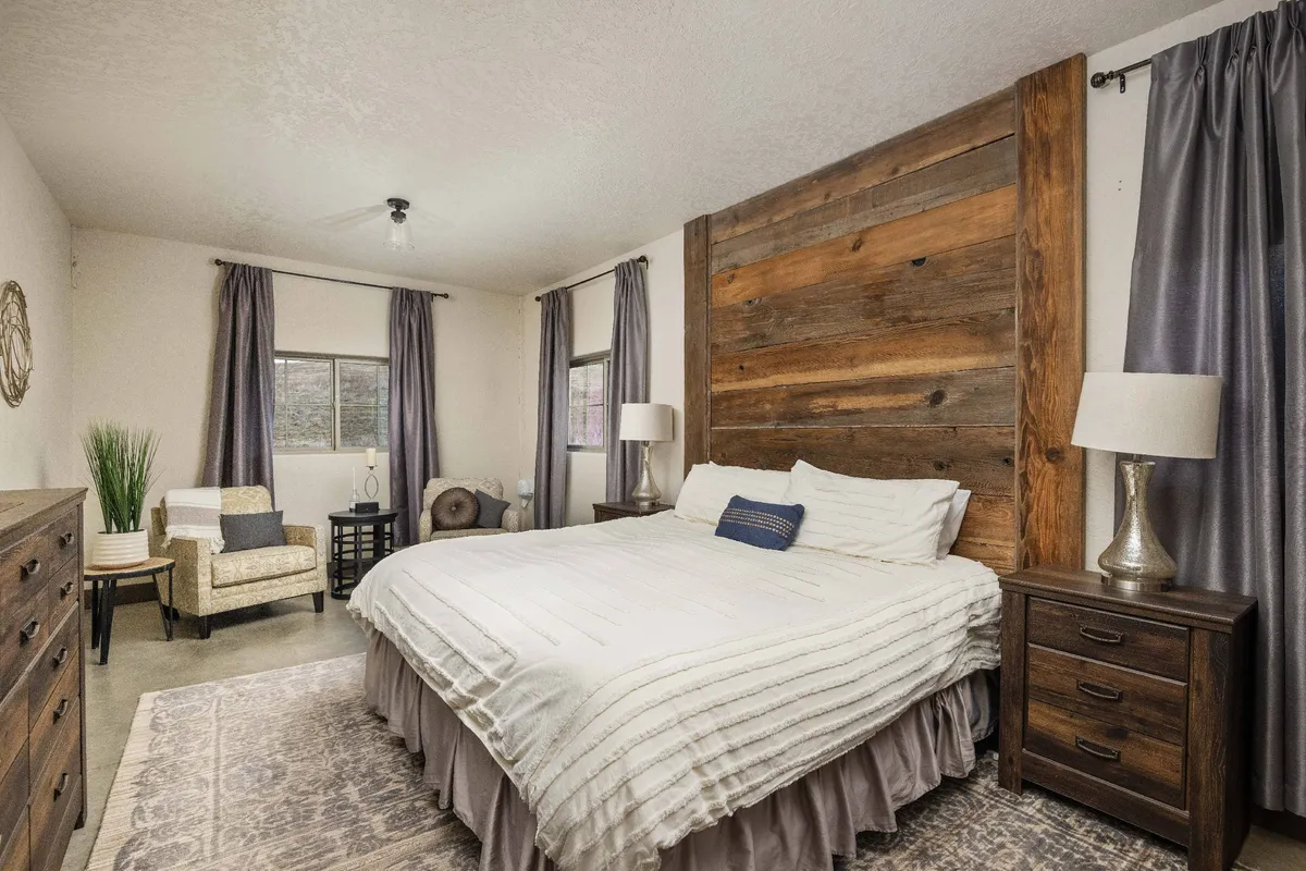 This image presents a warmly lit bedroom with a wood accent wall, a plush bed with high-quality bedding, nightstands, a comfortable chair, and a window allowing natural light to softly brighten the space.