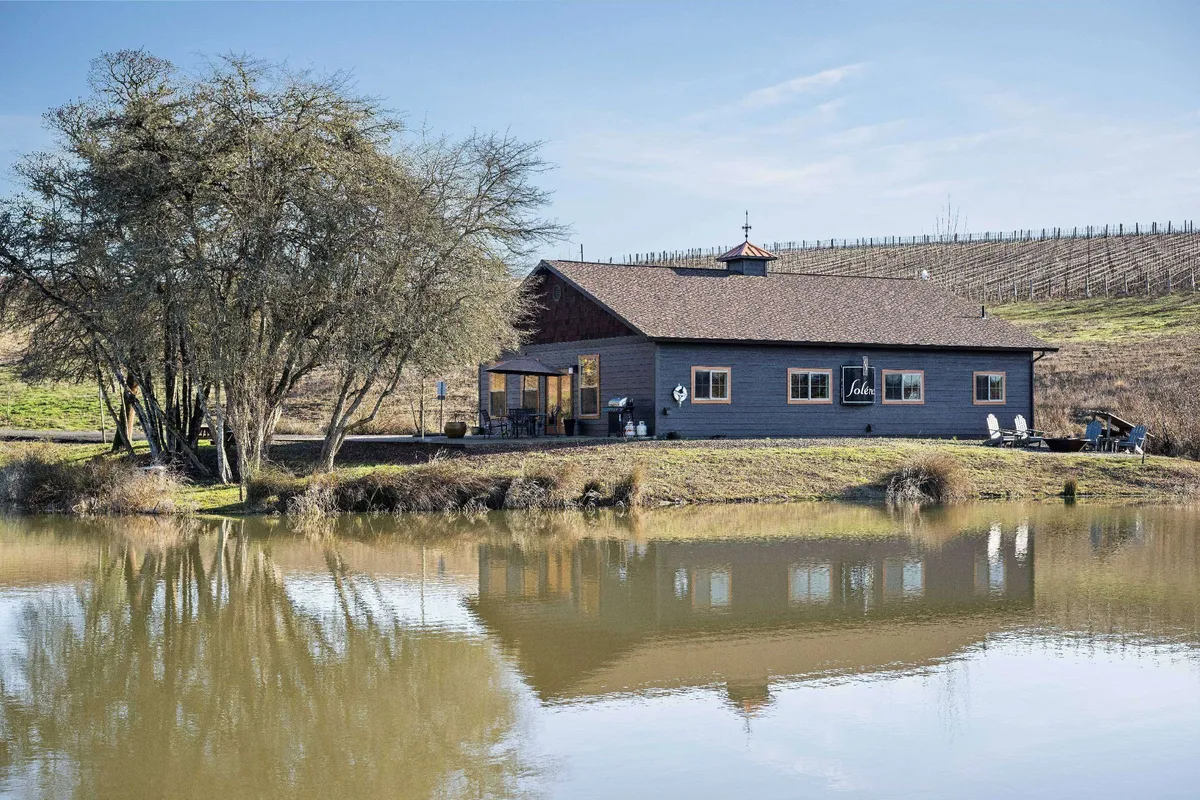 A peaceful exterior shot featuring the main house, the reflective pond, and surrounding trees. The image captures subtle changes in light, suggesting either an early morning mist or a tranquil sunset, evoking calm and introspection.